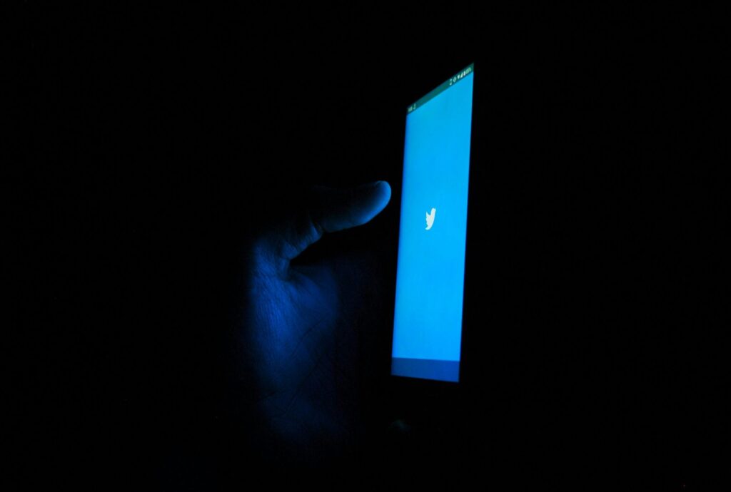 Picture of a hand holding a phone in the dark. The phone has an image of the old Twitter logo on it.
